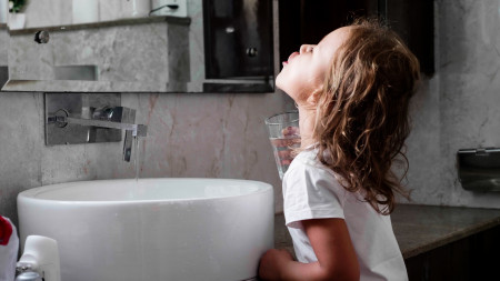Little curly child girl rinses her mouth with water in bathroom, side view.